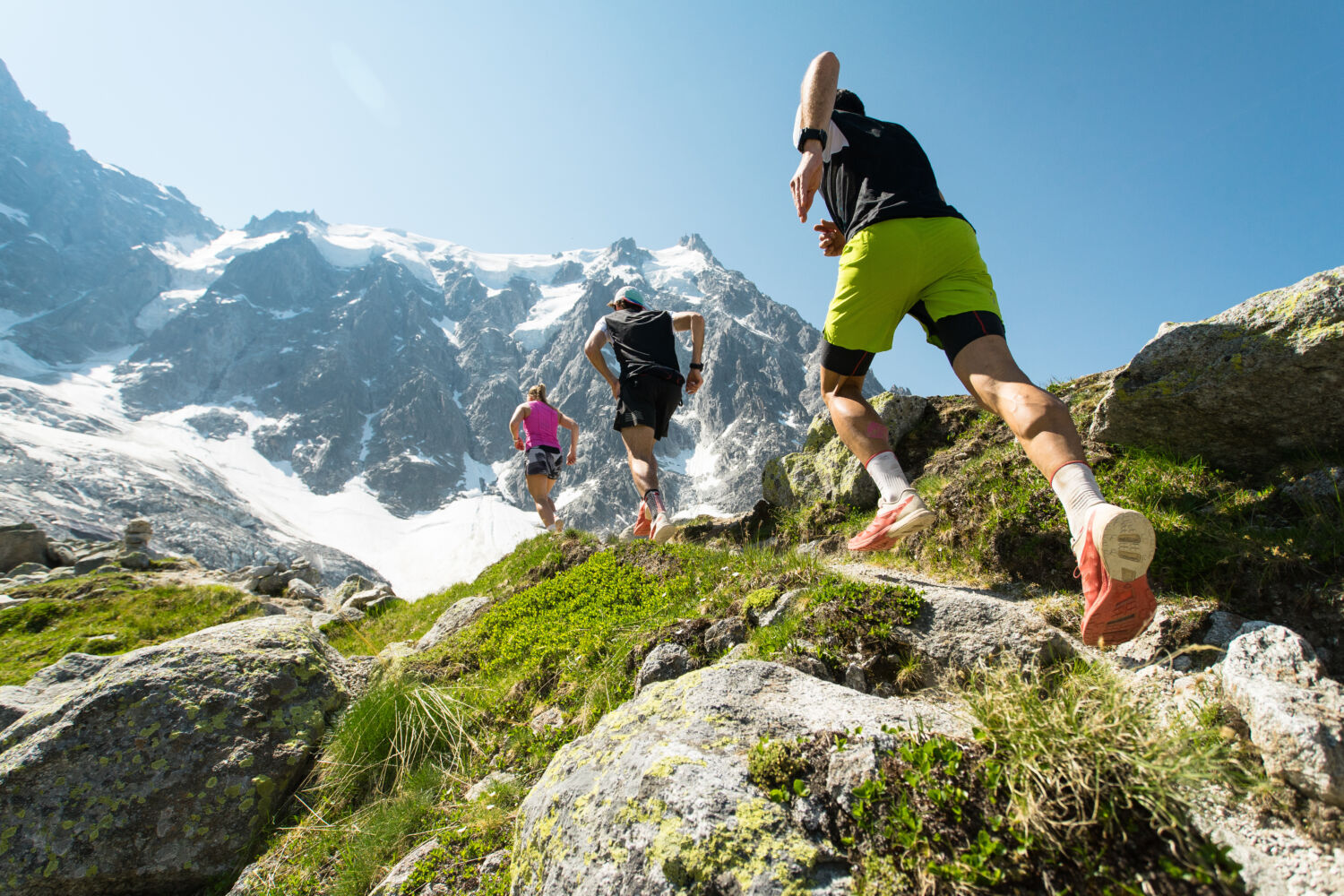 trail running dans les Alpes Trois coureurs de trail, deux hommes et une femme, courant sur un sentier escarpé dans les montagnes des Alpes par une chaude journée d'été ensoleillée.