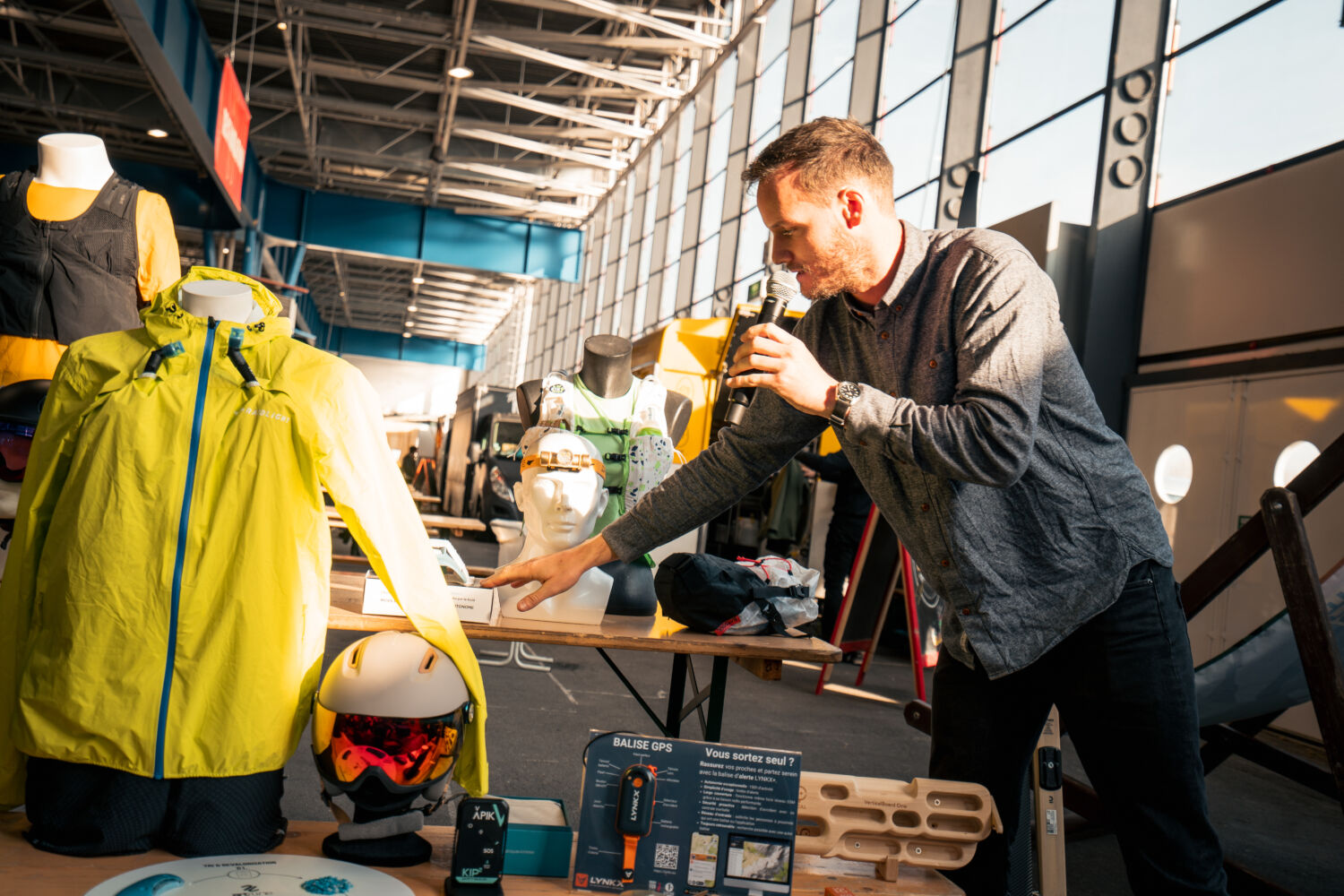 Sport Achat French Outdoor Awards Un homme présente du matériel de sport en plein air lors d'un salon, mettant en avant une veste imperméable, un casque de ski et un GPS de sécurité. L'événement se déroule dans un espace lumineux avec une ambiance professionnelle et dynamique.
